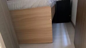 Een smalle gang met een houten vloer die leidt naar een kamer met een bed en een zwart meubelstuk.