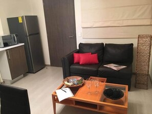 A compact living space with a kitchenette, a sofa with red cushions, a coffee table with glasses, and a woven basket.