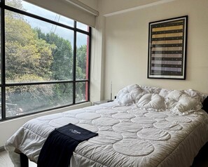 A bedroom with a large window, a bed with white bedding, and a framed artwork on the wall.