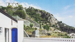 Un bâtiment blanc avec une porte bleue, un mur blanc orné d’une bordure décorative, et une vue sur une colline rocheuse couverte de végétation.