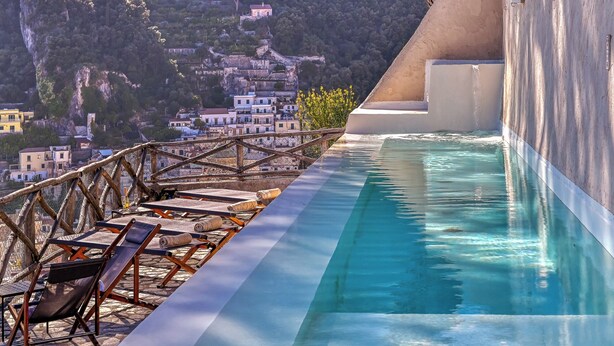 Une piscine à débordement située sur le toit, avec des chaises longues et une vue sur une ville perchée sur une colline.