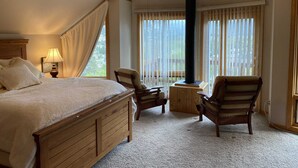 Ein Schlafzimmer mit einem hölzernen Bett, zwei Stühlen, einem kleinen Tisch, einem Deckenventilator und einem großen Fenster mit Vorhängen.