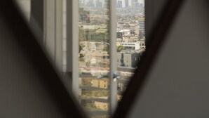 Vista pela janela com padrão geométrico, mostrando um horizonte urbano com edifícios e céu claro.