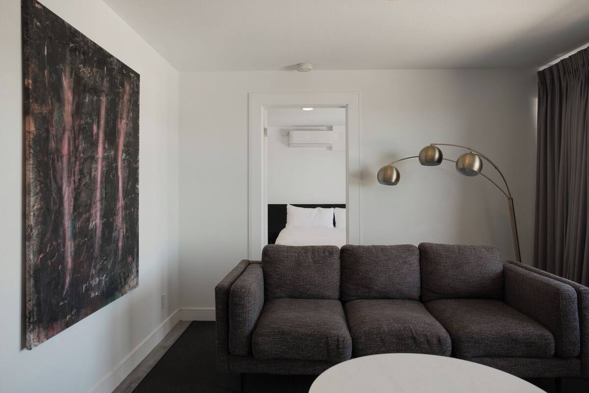 A modern hotel room with a grey sofa, a large abstract painting, a bedside table, and a floor lamp.