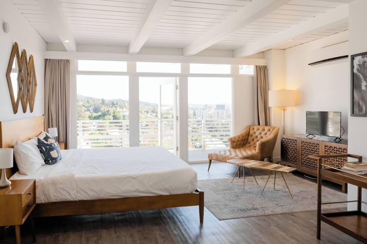 A hotel room with a large bed, a sitting area with a chair, a TV, and a view of the city.