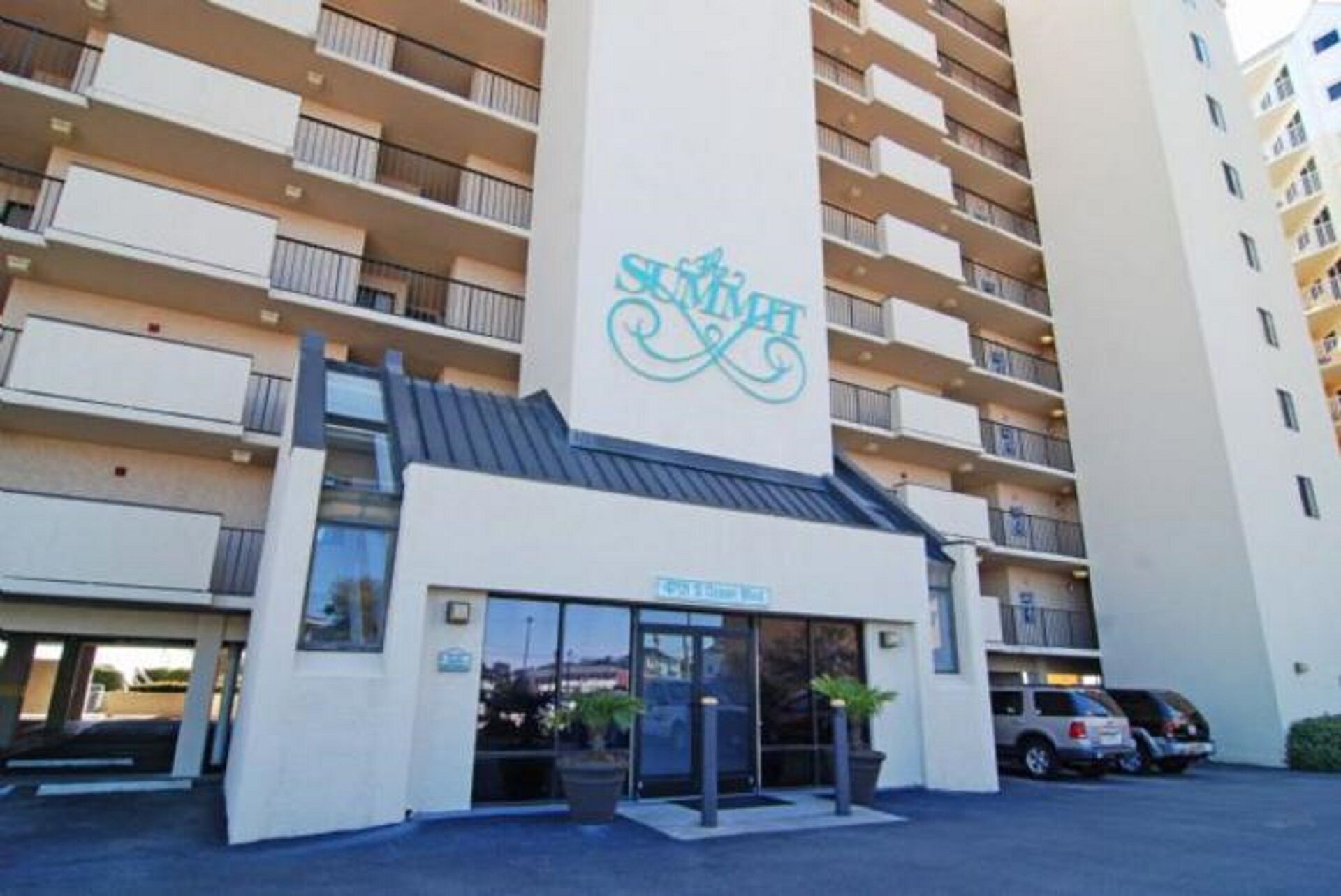 A modern hotel with a central entrance, multiple balconies, and a parking area.