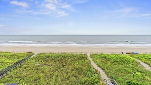 A beach with a wooden boardwalk leading to the water, grassy areas, and a clear sky.