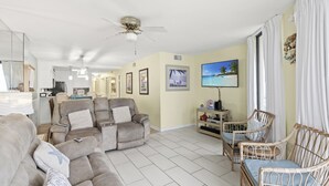 A spacious living room with a beige sofa, a reclining chair, and wicker chairs. There is a ceiling fan, a flat-screen TV, and framed pictures on the walls.