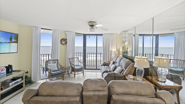 A spacious living room with a large sofa, two armchairs, a coffee table, and a television. The room has large windows with a view of the ocean.