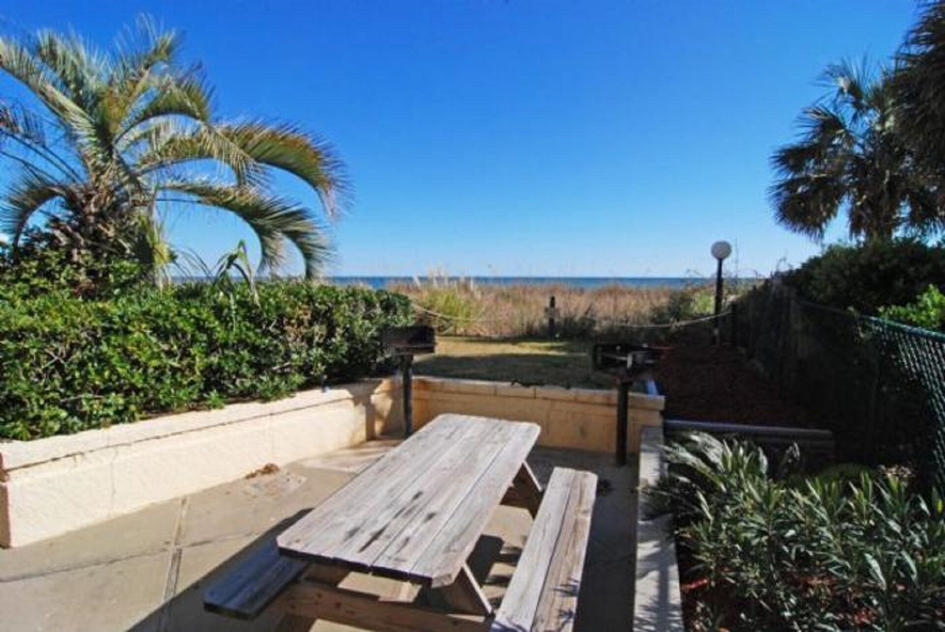 Zona de pati amb taula i bancs de fusta, envoltada de verdura i vista a la platja.