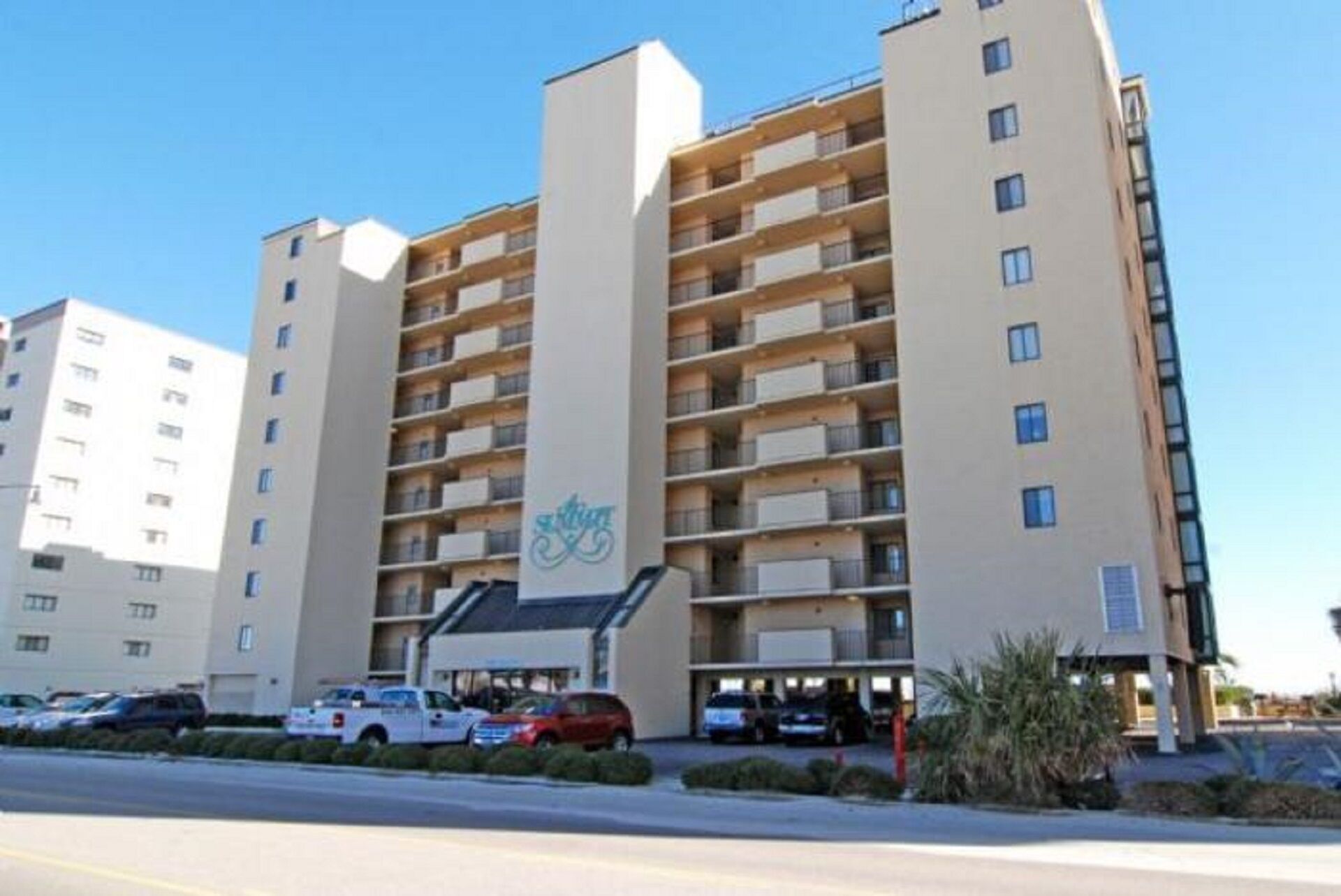A multi-story hotel with a central entrance, balconies on each floor, and a parking area in front.