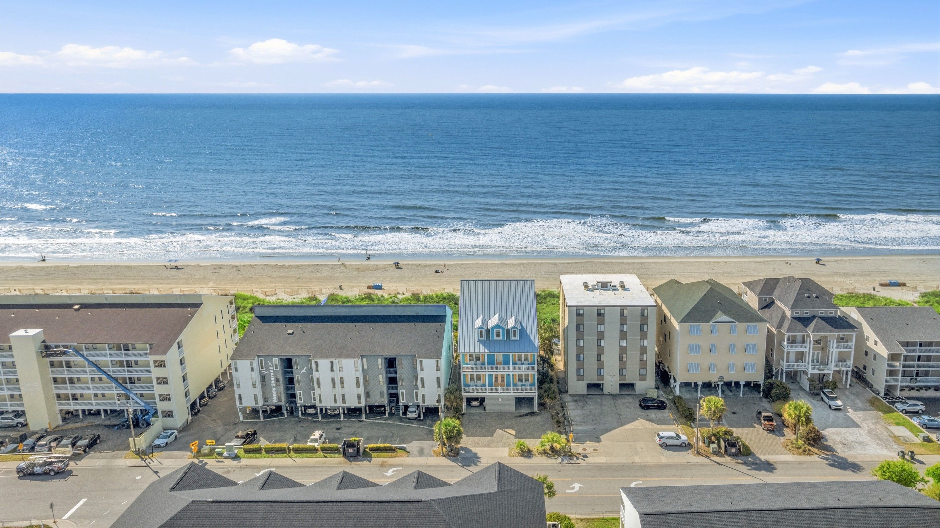 Et kysthotel med flere bygninger, en parkeringsplads og en strand i baggrunden.
