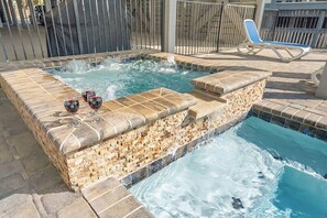 A hot tub with a stone mosaic design, surrounded by a pool and a lounge chair.