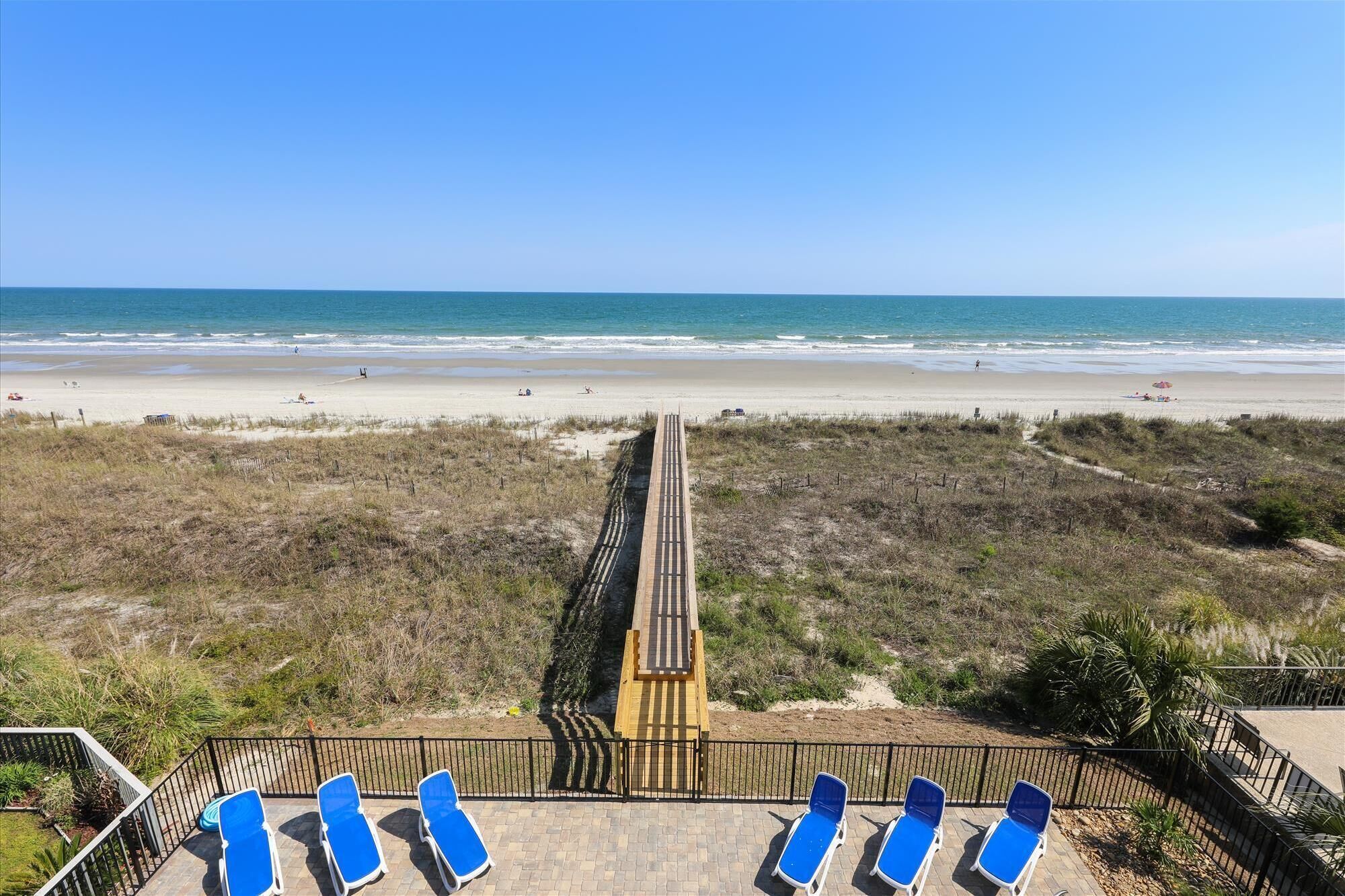 A beach view with a wooden boardwalk leading to the shore, and a row of blue lounge chairs on a patio area.
