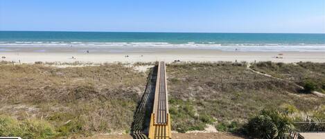 A beach view with a wooden boardwalk leading to the shore, and a row of blue lounge chairs on a patio area.