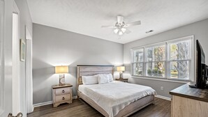 Une chambre à coucher comprenant un lit, des tables de chevet, un ventilateur de plafond, une fenêtre avec des stores et un téléviseur posé sur une commode en bois.