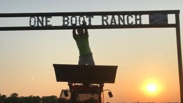 A person standing on a tractor at a gate labeled "ONE BOOT RANCH" during sunset. - Tiny House Living on the ranch in East Texas (Point)