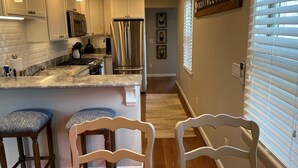 A modern kitchen with white cabinets, stainless steel appliances, and a marble countertop. There are two bar stools with patterned cushions. - Amazing WEST END location, steps away from all the action! (Provincetown)