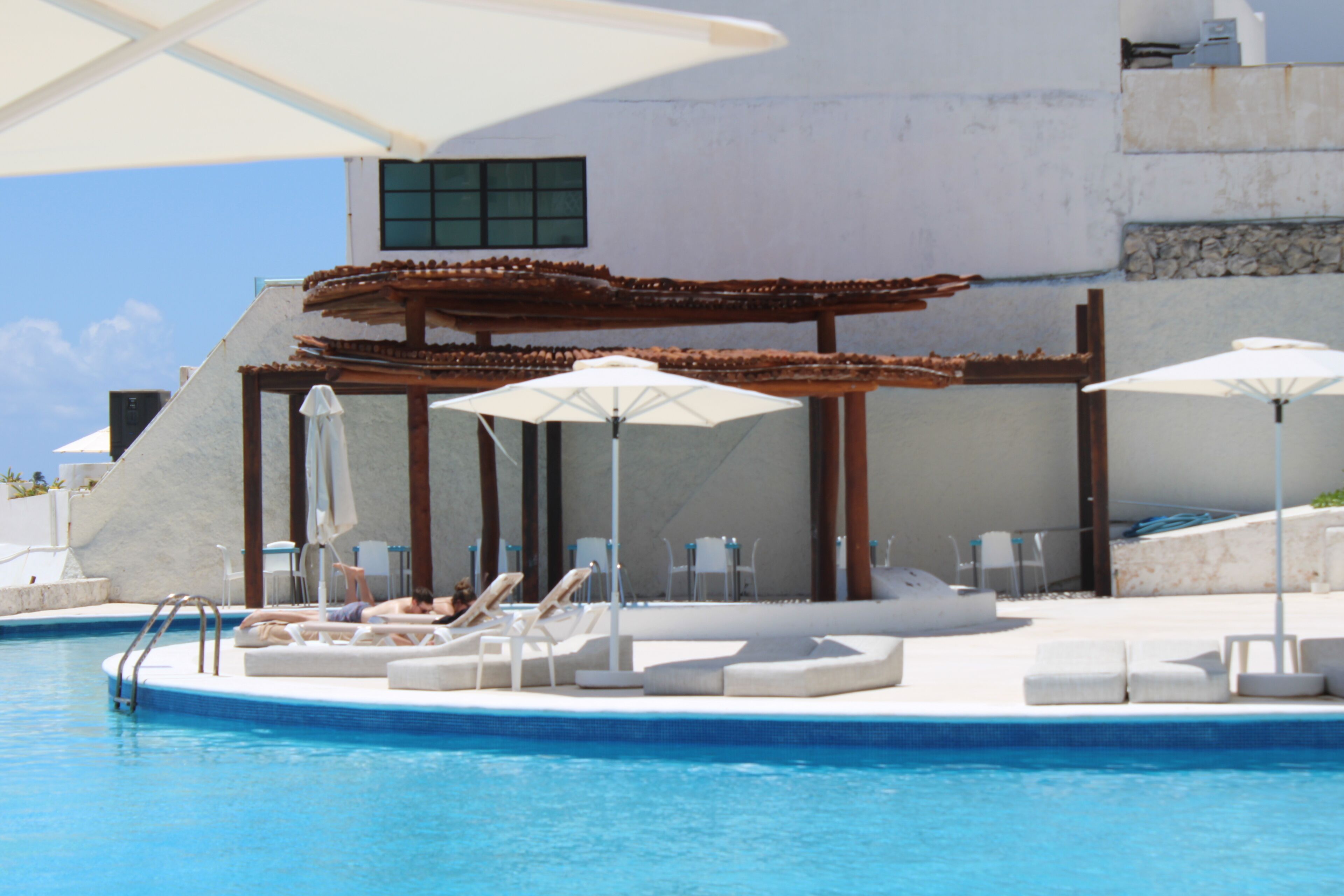 Zona de piscina con sillones de descanso y sombrillas, una pérgola y un edificio con fachada de piedra.