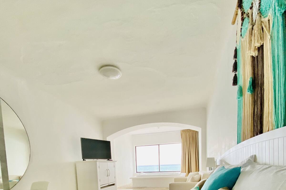 A hotel room with a large bed, a flat-screen TV, a desk, and a window with a view of the sea.