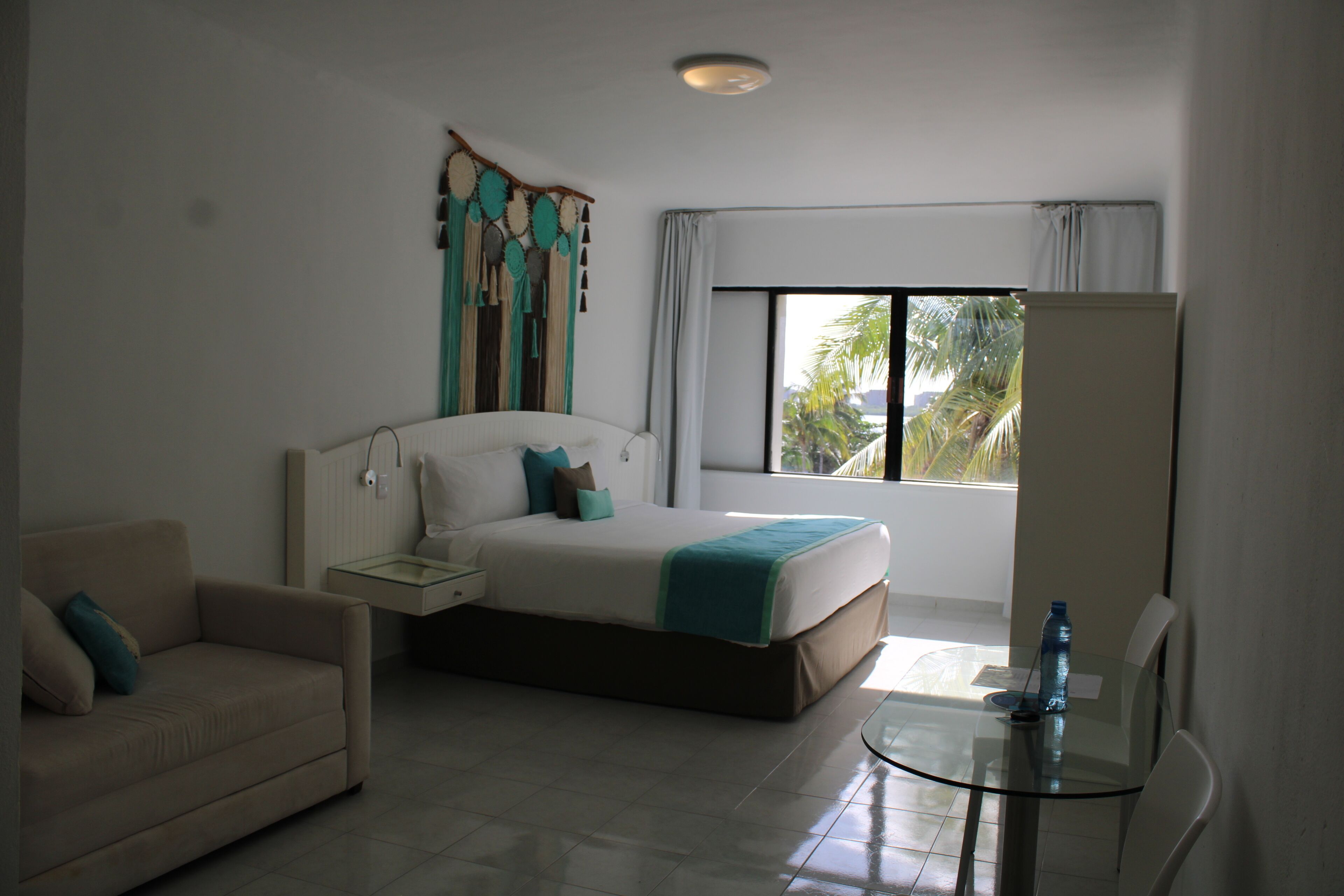 Habitación de hotel con cama, sofá, mesa de vidrio y ventana con vista a palmeras.