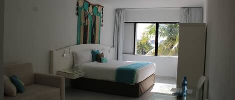 Habitación de hotel con cama, sofá, mesa de vidrio y ventana con vista a palmeras.