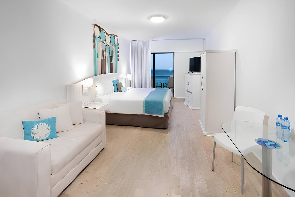 A modern hotel room with a bed, a sofa, a glass table with water bottles, a TV, and a view of the sea.