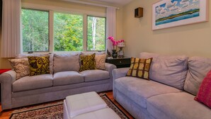 A living room with two sofas, a coffee table, and a large window with curtains. - Theatre in the Wood Too! - I’m the fancy sister (Stowe)