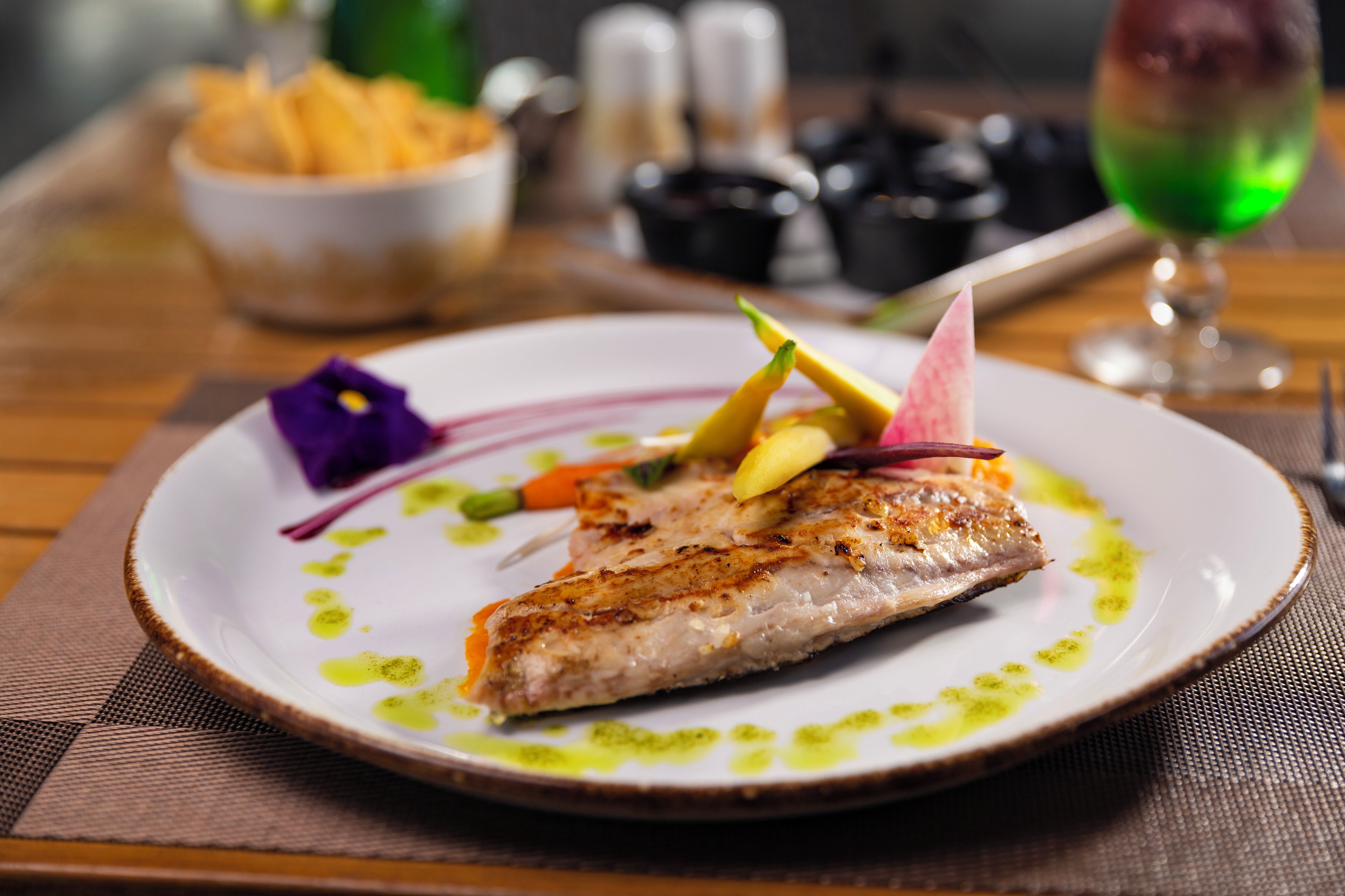 Ein Teller mit gegrilltem Fisch, garniert mit Gemüse und einer violetten Blüte, auf einer strukturierten Unterlage.