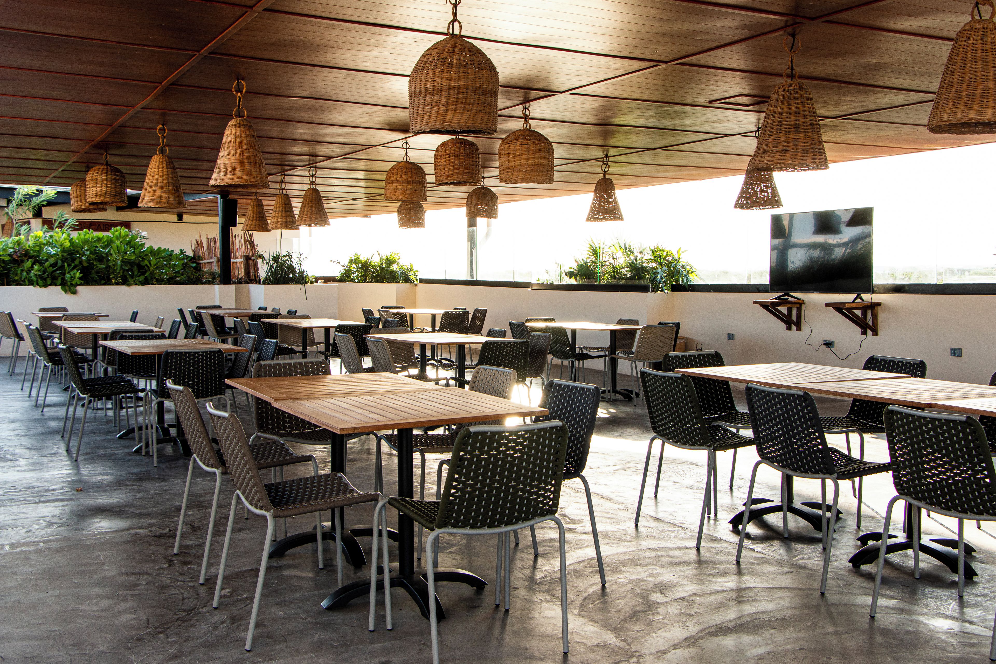 An indoor dining area with multiple tables and chairs, hanging wicker lamps, and a large flat-screen TV mounted on the wall.