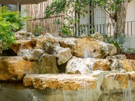 Un jardín de rocas con una pequeña cascada, rodeado de abundante vegetación y una valla de bambú.
