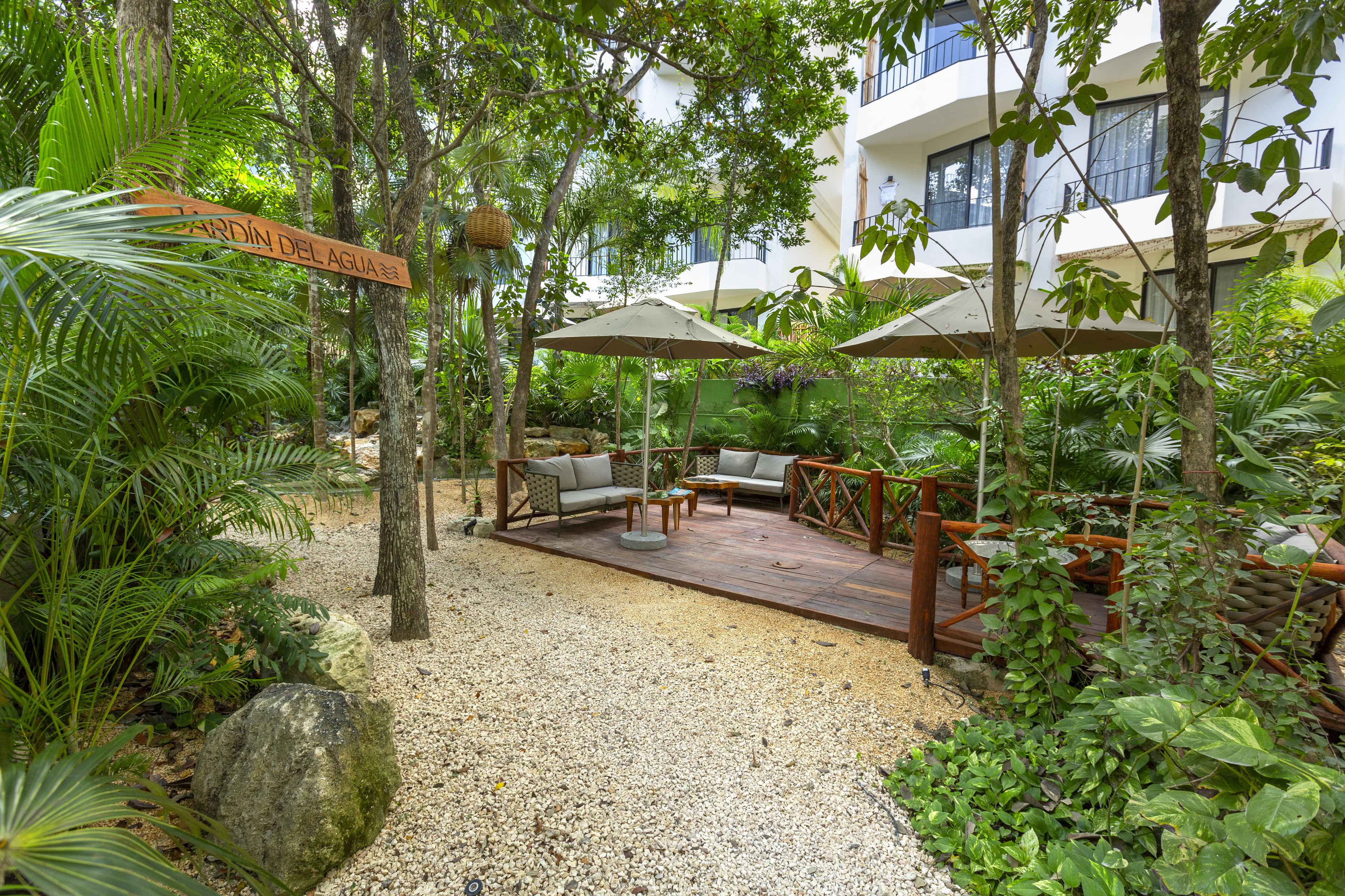 A tropical garden patio with a wooden deck, outdoor furniture, and a sign reading 'Garden Del Agua'.