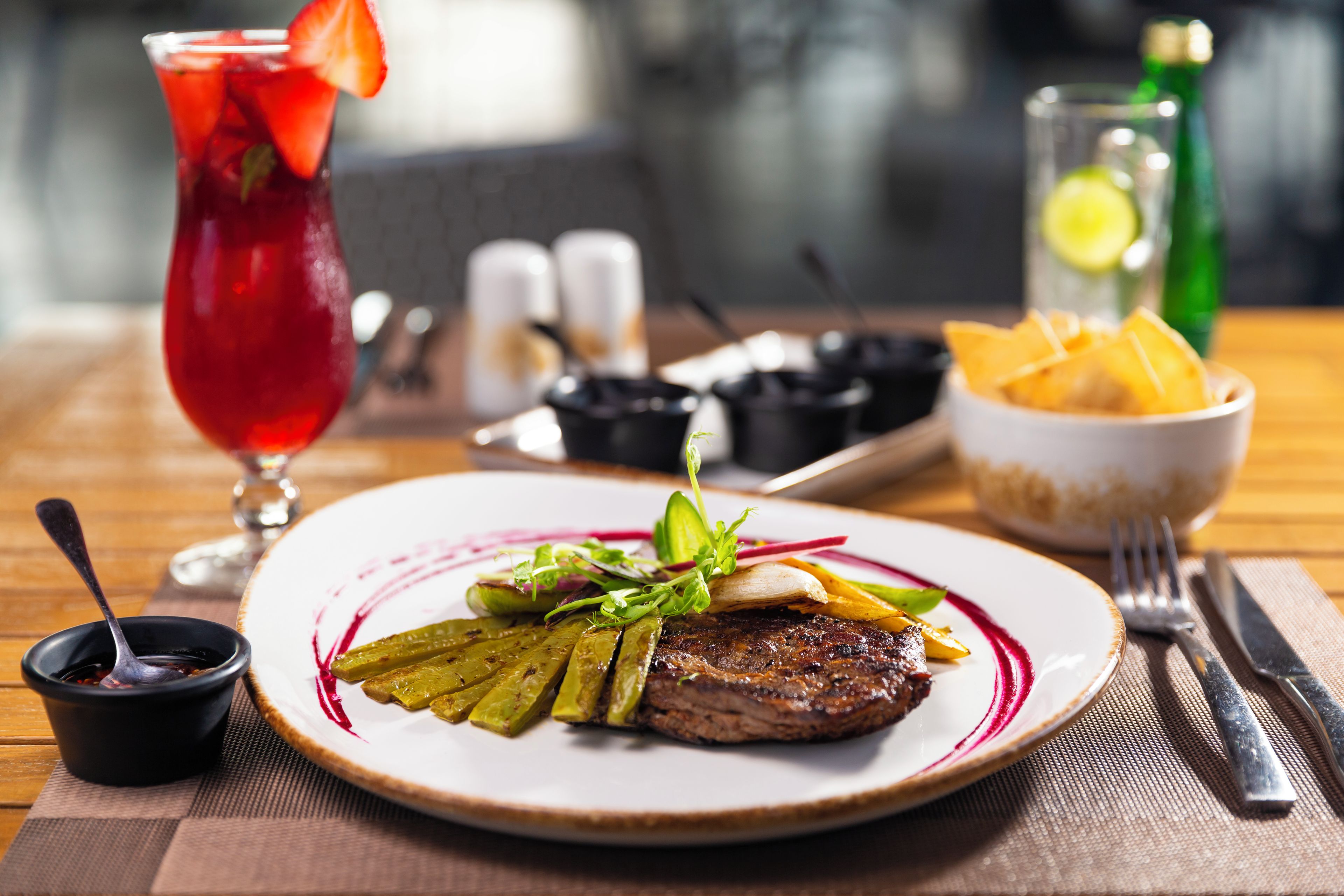 Un plato de comida con bistec, pepinillos, verduras, un vaso de bebida roja y un tazón de papas fritas sobre una mesa.