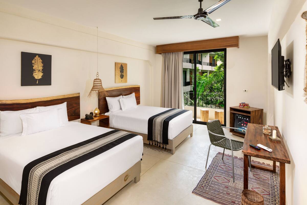 A hotel room with two beds, a TV, a desk, and a balcony with a view of greenery.