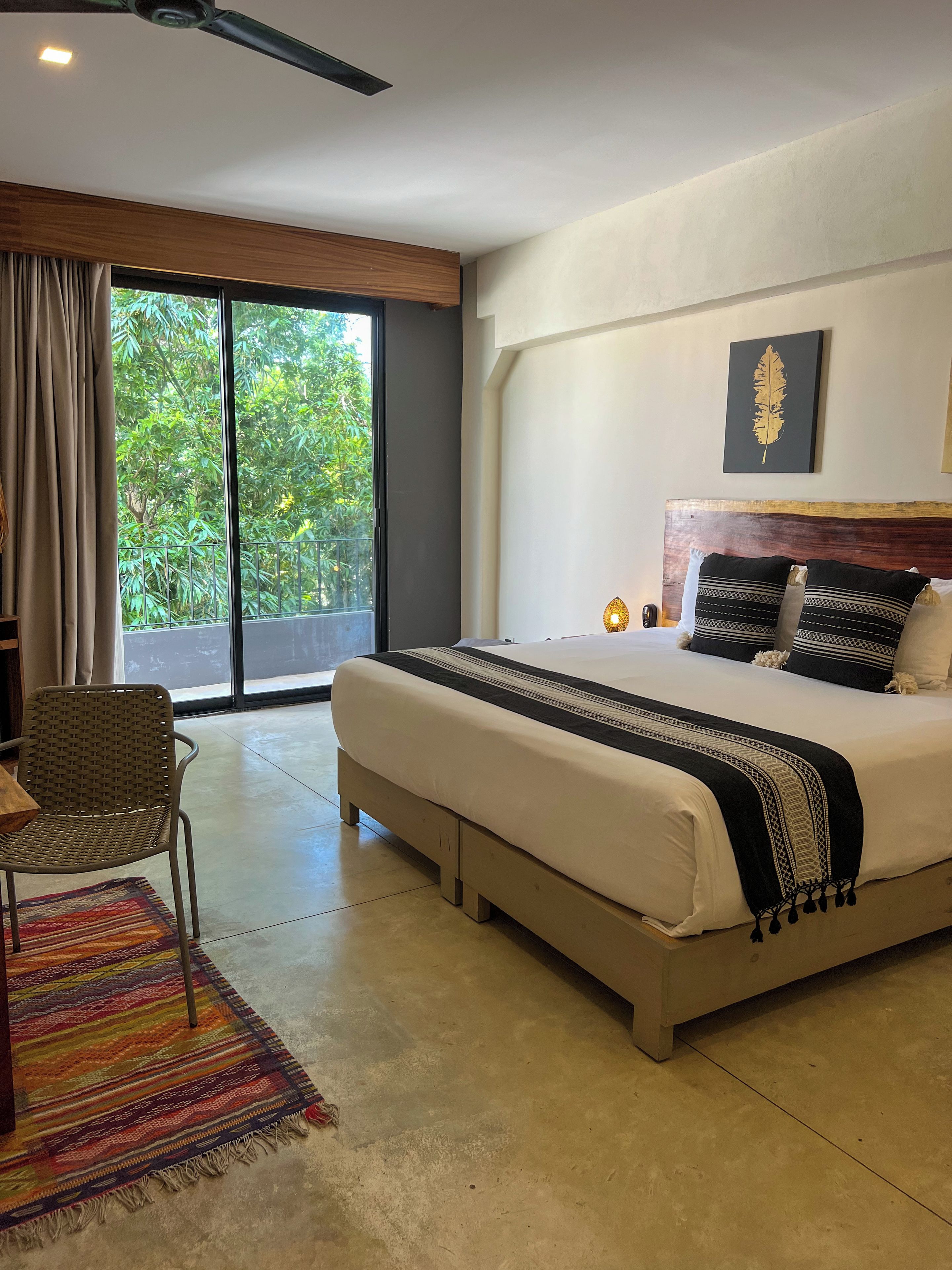 A modern hotel room with a large bed, a desk, and a view of greenery through the window.