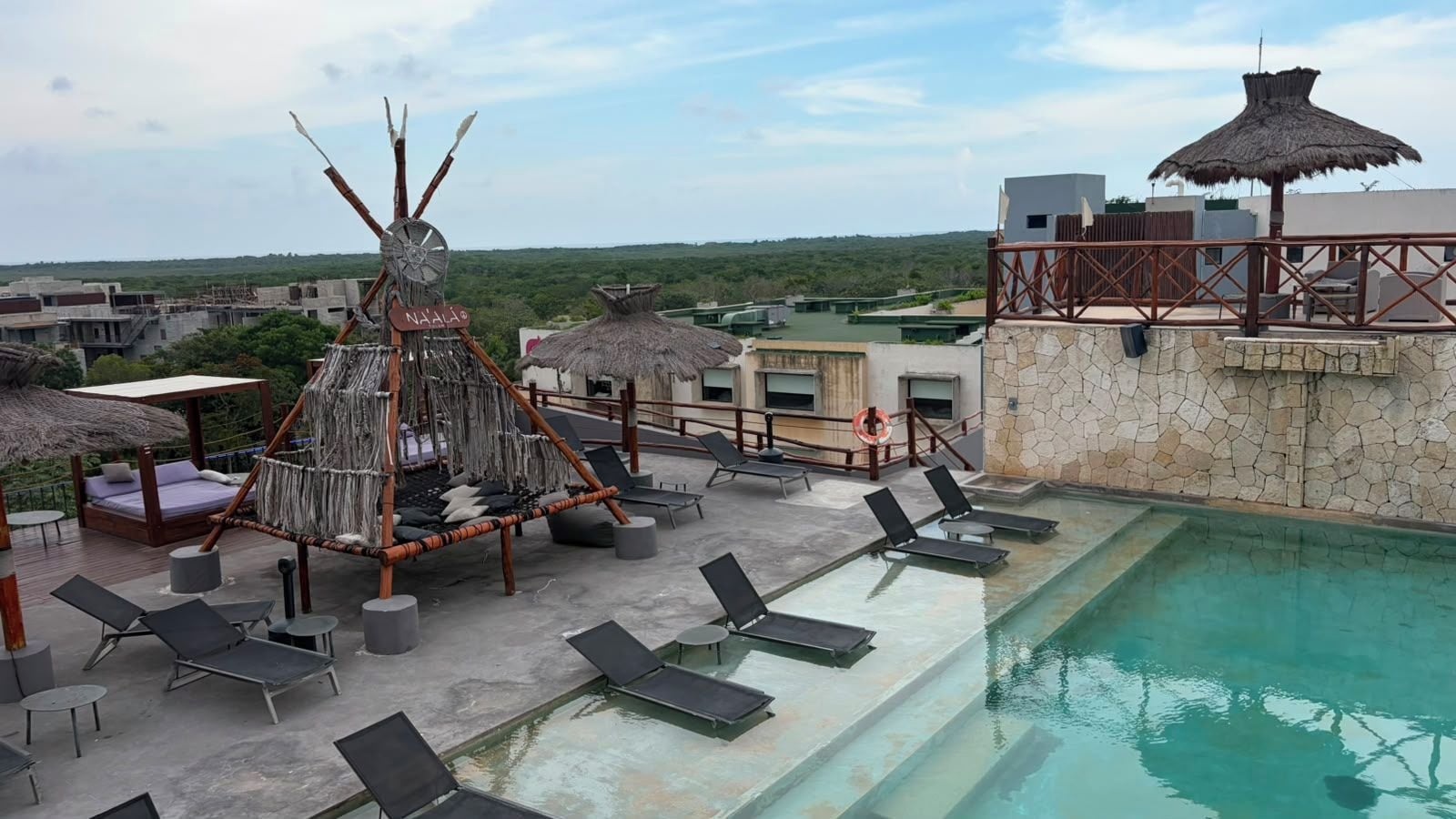 A rooftop pool area with lounge chairs, a thatched-roof seating area, and a view of the surrounding landscape.