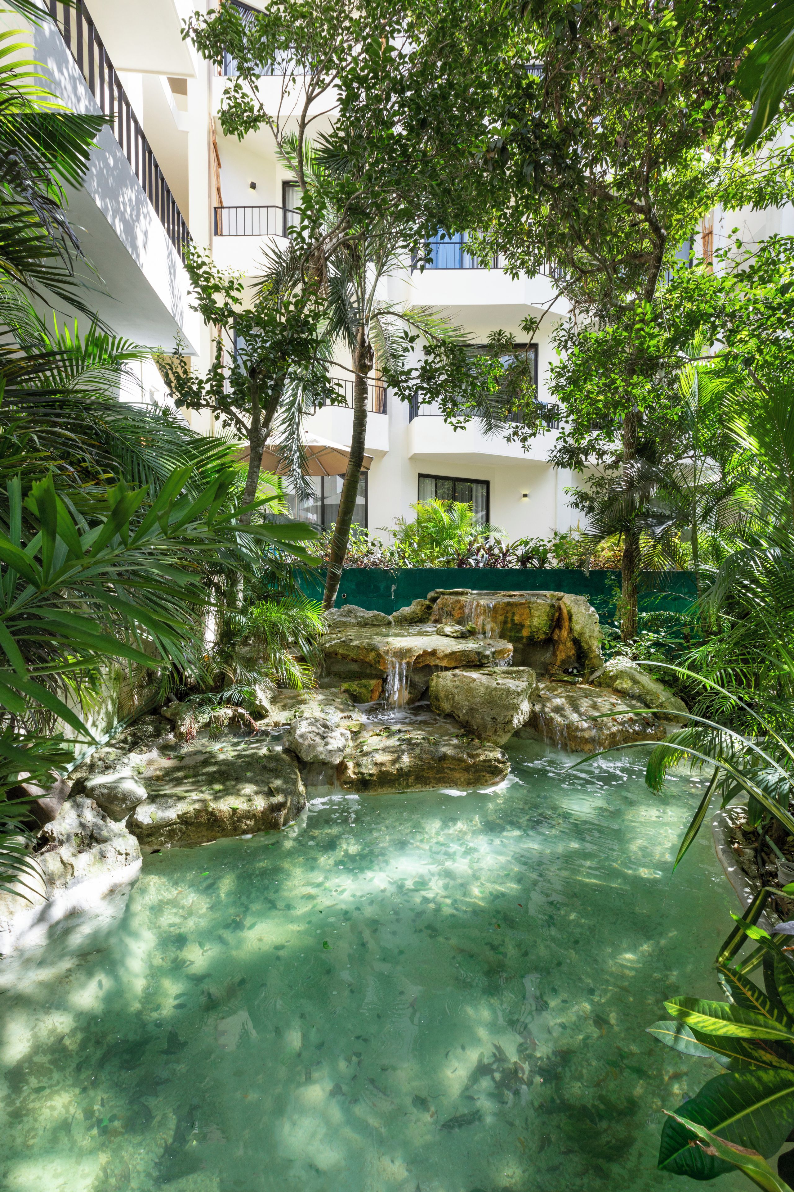 A clear water feature with rocks and a waterfall, surrounded by lush greenery and a building with balconies.