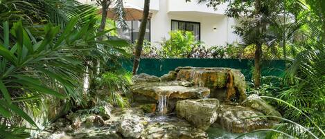 Un elemento acuático con agua cristalina, rocas y una cascada, rodeado de abundante vegetación y un edificio con balcones.