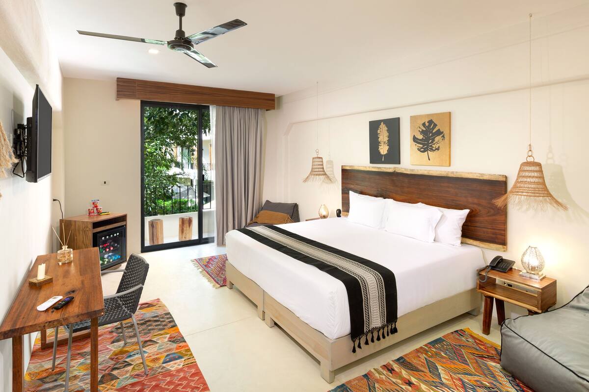 A modern hotel room with a large bed, a desk, a chair, a TV, a balcony with a view of greenery, and a ceiling fan.