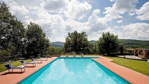 A rectangular swimming pool with clear blue water, surrounded by a brick patio. There are lounge chairs along the pool's edge, and the area is landscaped with trees and grass. - Colombaio Apartment (Pietraviva)