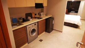 A compact hotel room with a kitchenette, a washing machine, a compact refrigerator, and a bed with a white duvet.