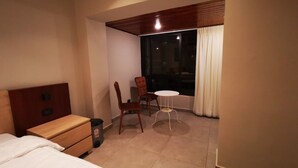 A hotel room with a bed, a wooden nightstand, a chair, a small round table, and a window with curtains.