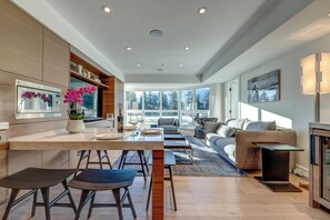 A modern kitchen with a central island, bar stools, and a dining table. There is a sofa and a coffee table in the living area, with a large window providing a view of the outdoors.
