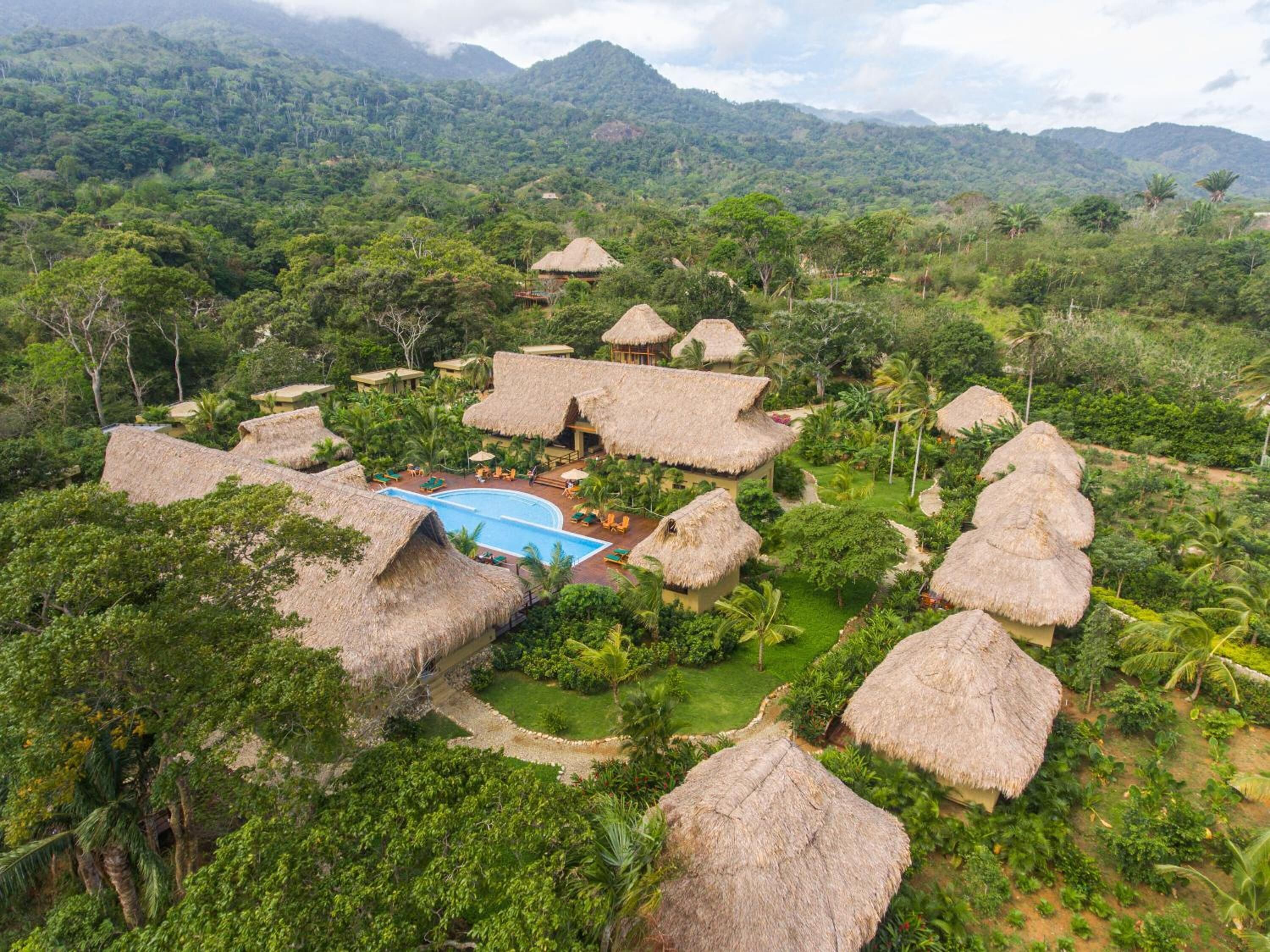A resort with thatched-roof huts, a swimming pool, and lush greenery.