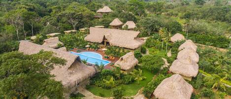 A resort with thatched-roof huts, a swimming pool, and lush greenery.