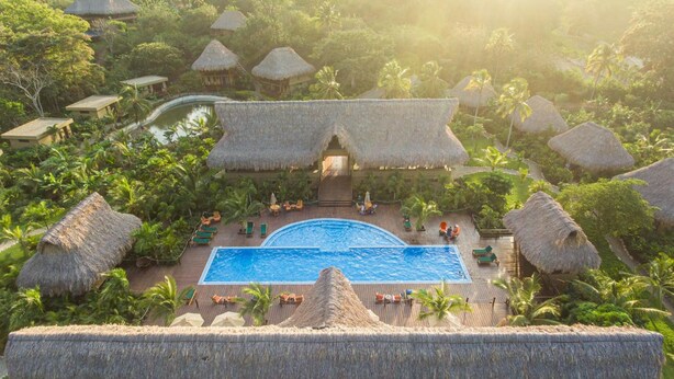 Um resort com piscina central rodeada por estruturas com telhados de palha e vegetação exuberante.