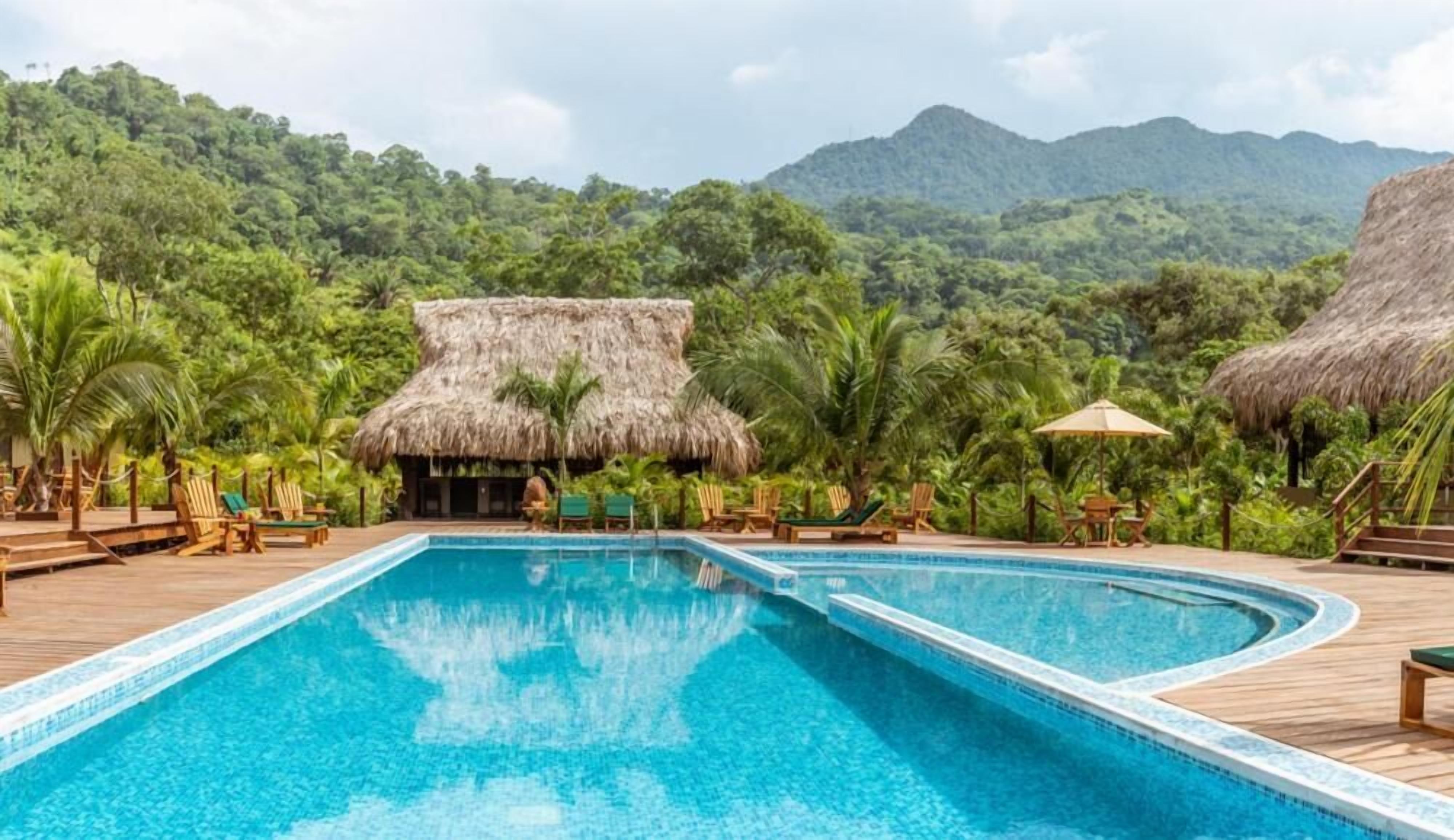A pool area with a thatched-roof structure, surrounded by lounge chairs and palm trees, set against a backdrop of lush green hills.