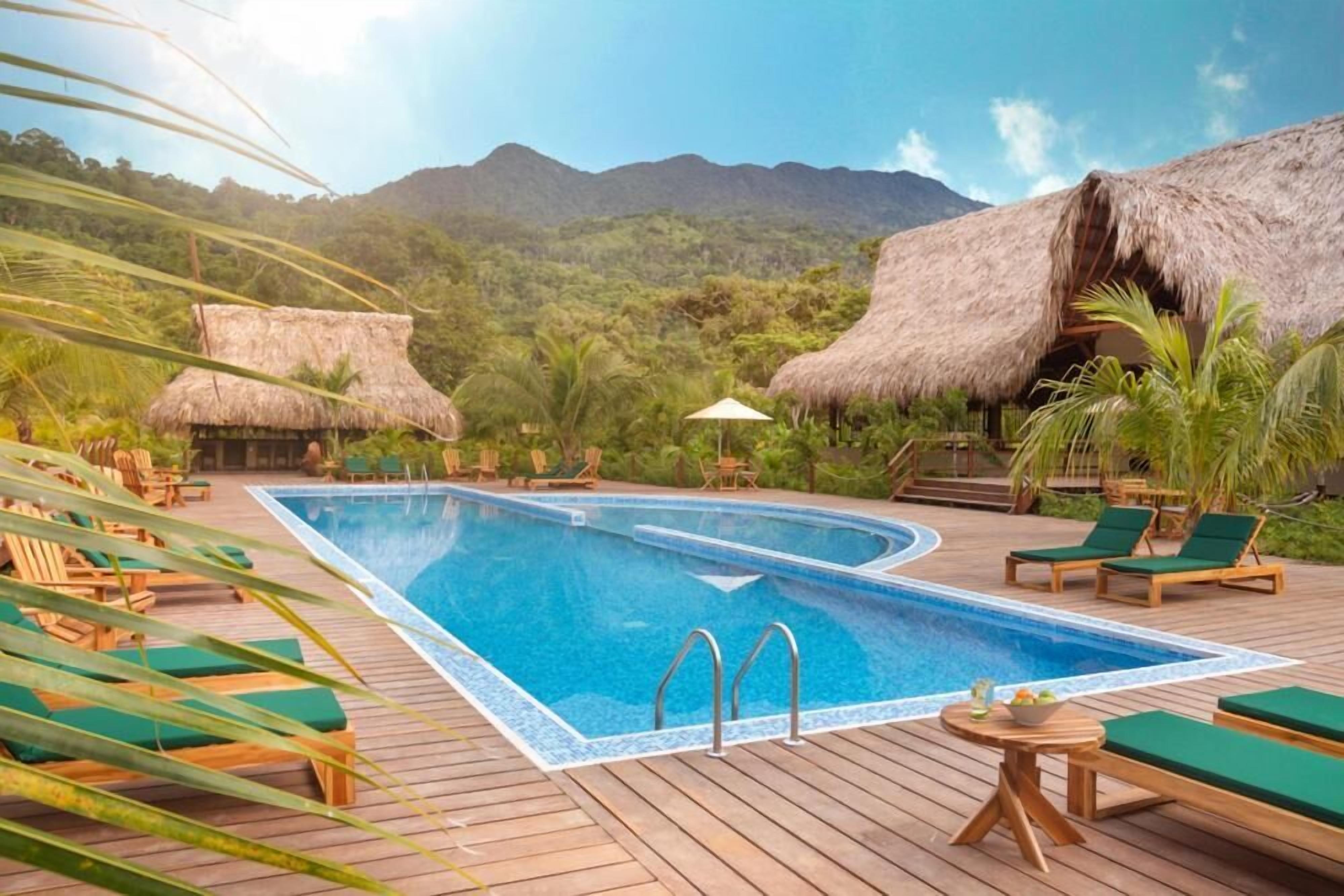 Un espace piscine offrant une vue dégagée sur les montagnes, avec une terrasse en bois et des constructions à toits de chaume.
