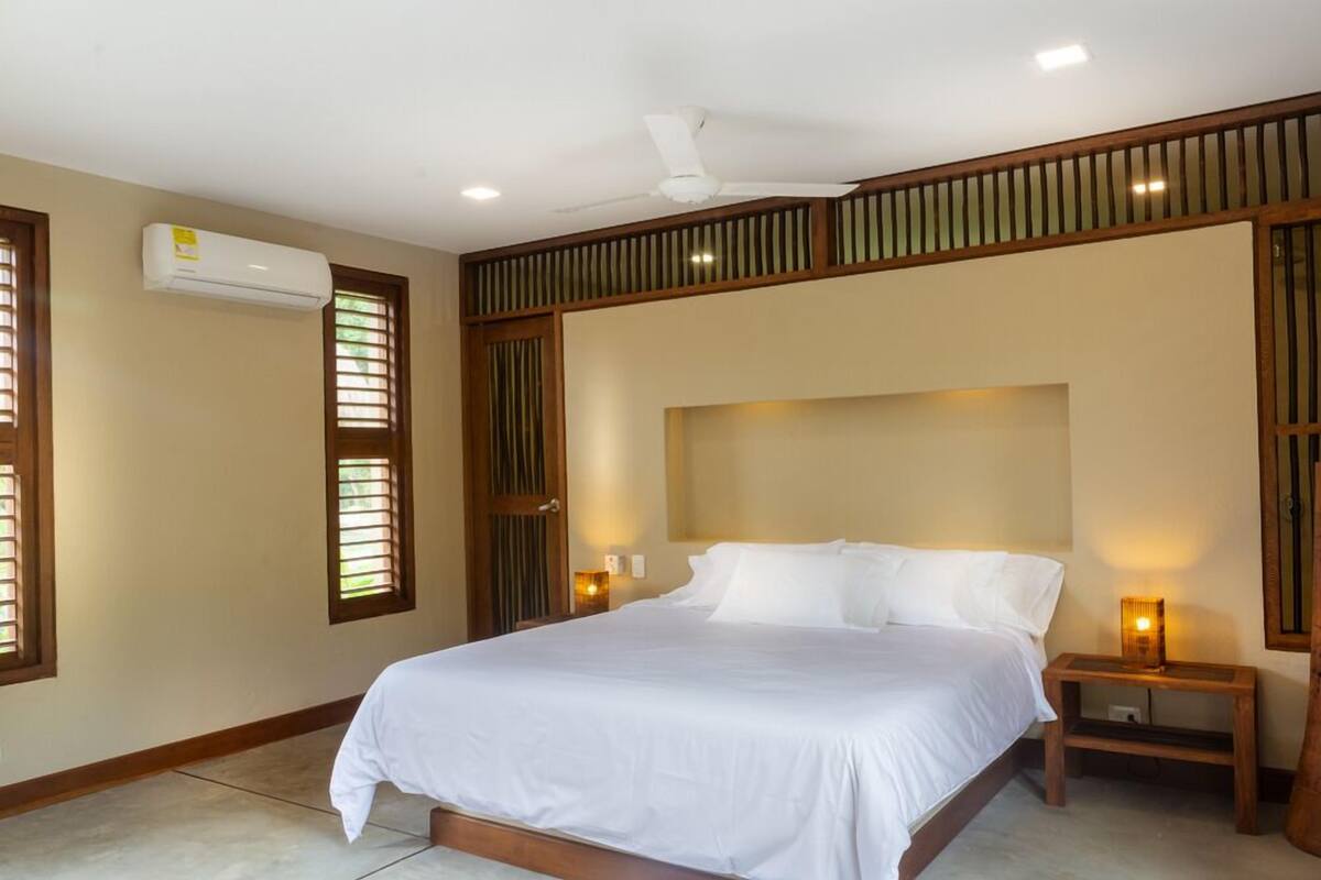 A bedroom with a bed, an air conditioning unit, a ceiling fan, and two bedside lamps.