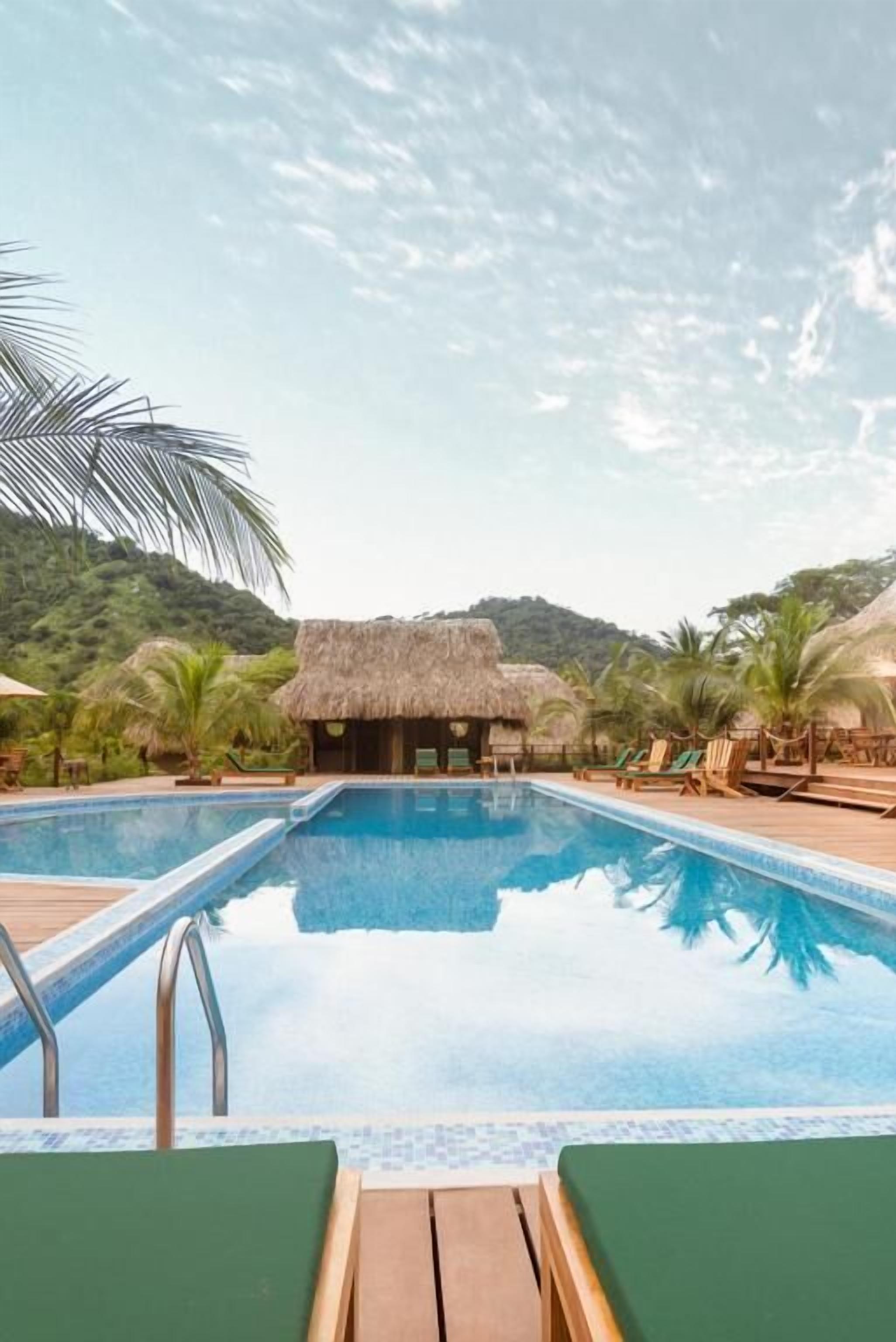 Una piscina con sdraio e un padiglione con tetto di paglia, circondata da palme e un paesaggio collinare.